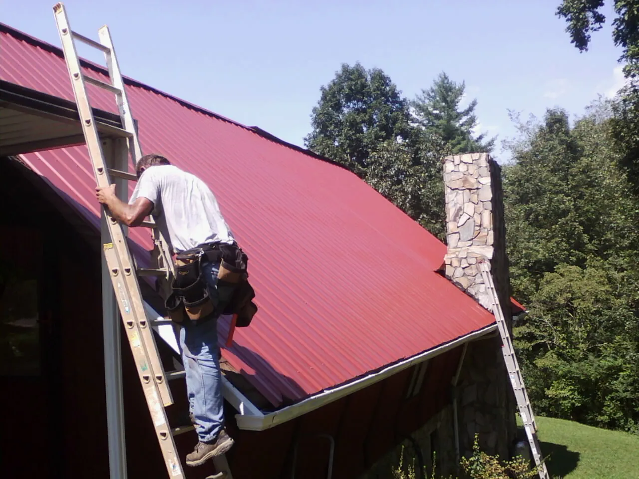 Roofer on a ladder beside a red metal roof and stone chimney — Soffit & Fascia Repair by Roofing Brothers in Geff